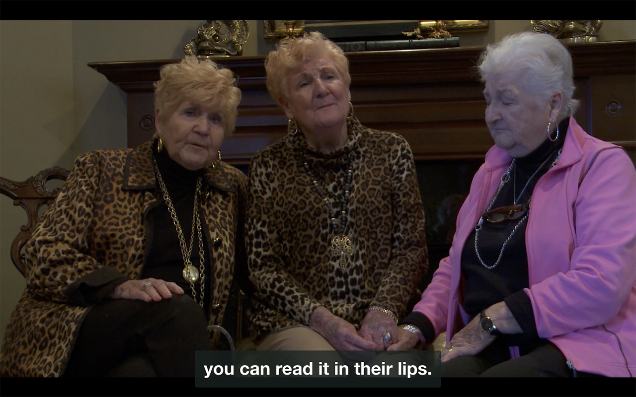 Three elderly white women sit next to each other; two of them wear leopard-print and have matching strawberry blonde hair. The third woman wears a pink track jacket and closes her eyes, as she holds the hand of the woman in the center. They all appear to be singing. A caption at the bottom of the frame reads: “you can read it in their lips.